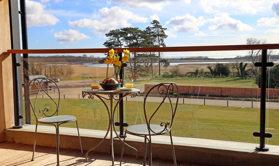 View from a balcony, two bistro chairs and a table with flowers and food and drink, in the background a beautiful view of fenland and a summer sky.