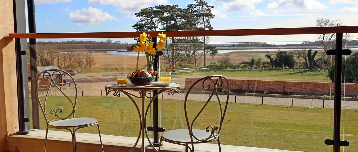View from a balcony, two bistro chairs and a table with flowers and food and drink, in the background a beautiful view of fenland and a summer sky.