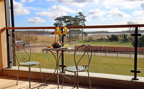 View from a balcony, two bistro chairs and a table with flowers and food and drink, in the background a beautiful view of fenland and a summer sky.