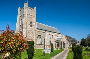Small village church with a churchyard and trees, under a blue sky.