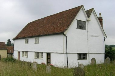 Rustic white two-storey building with red tiled roof. Gravestones outside in long grass.