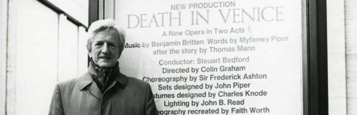 Peter Pears as an older man with white hair, in a 3/4 length coat, standing in front of MET sign