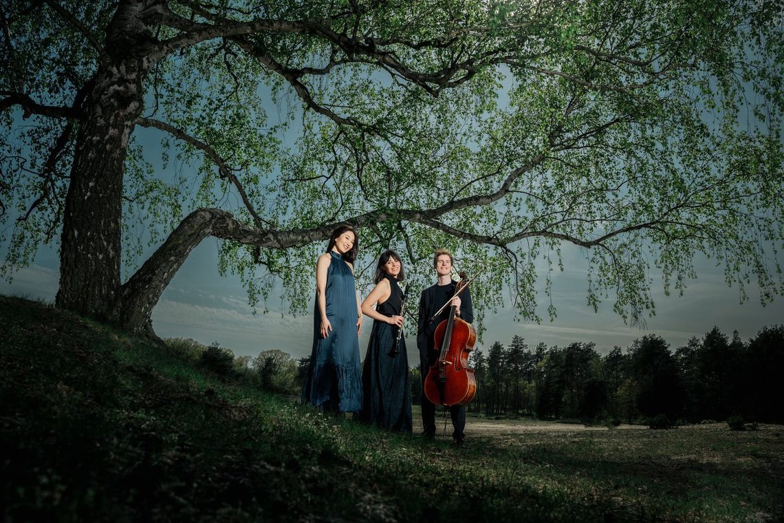 Man and two women wearing evening dress, standing in woodland with their string instruments