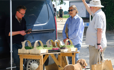 A man stood in front of a navy van with a large awning coming off the side. Underneath is a table with plants on, and looking at the table is a blonde women wearing sunglasses and a man with a white hat, holding a bag, and their dog.