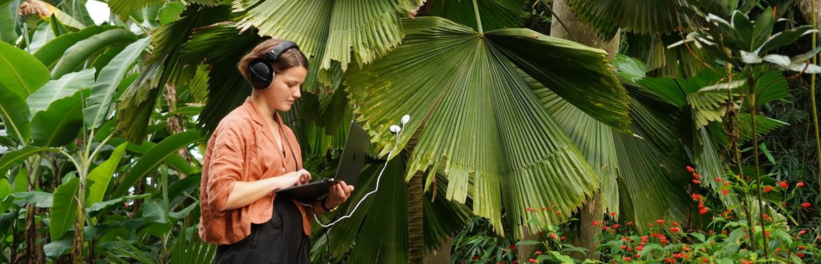 Young woman with brown hair wearing headphones and holding a laptop in the middle of a forest.