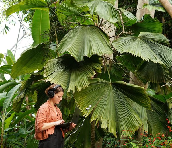 Young woman with brown hair wearing headphones and holding a laptop in the middle of a forest.
