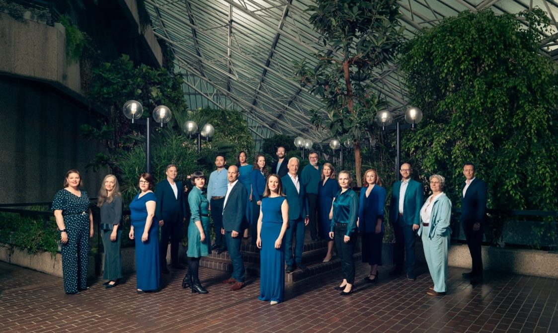 Group of men and women wearing blue and green clothing standing together under a large glass roof..