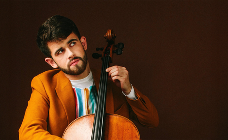 Hugh Mackay, man with short dark hair and beard wearing a brown jacket and holding a cello.