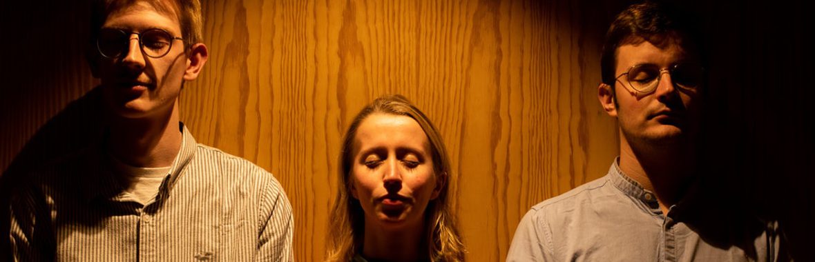 A blonde woman wearing a pink sweater stands inbetween two men wearing shirts, they all face forward with closed eyes.