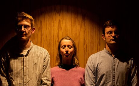 A blonde woman wearing a pink sweater stands inbetween two men wearing shirts, they all face forward with closed eyes.