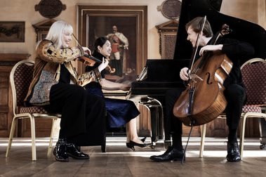 Three musicians playing piano and string instruments onstage and smiling.