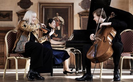 Three musicians playing piano and string instruments onstage and smiling.