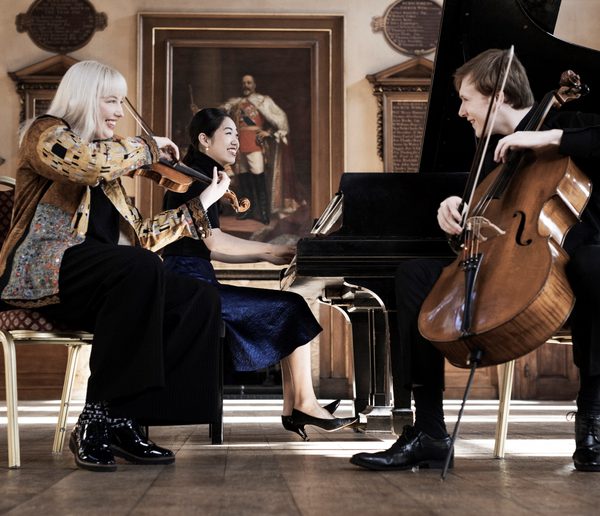 Three musicians playing piano and string instruments onstage and smiling.
