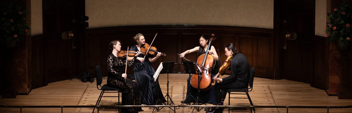 Four musicians sitting in the centre of the stage all wearing black, playing string instruments.
