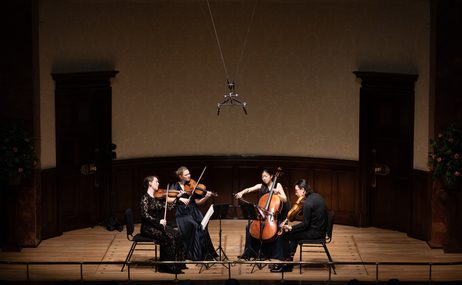 Four musicians sitting in the centre of the stage all wearing black, playing string instruments.