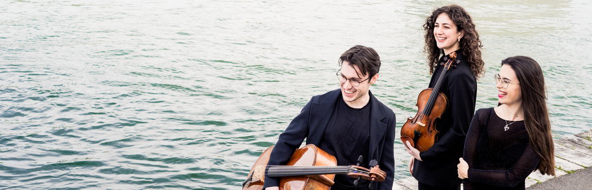 One male and two female musicians standing together holding musical instruments, with water in the background.