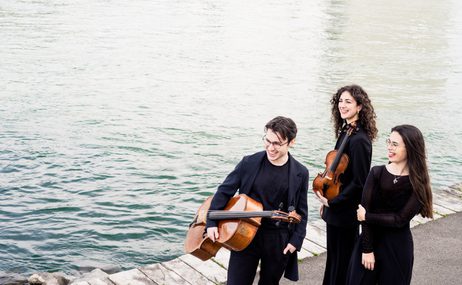 One male and two female musicians standing together holding musical instruments, with water in the background.