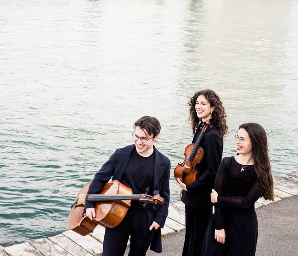 One male and two female musicians standing together holding musical instruments, with water in the background.