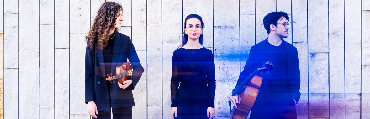 Trio Archai, one man and two women, all wearing dark clothing and holding musical instruments against a stone wall.