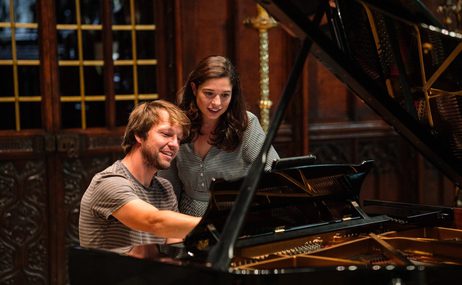Man with short brown hair and woman with long dark hair standing together at a piano looking at music.