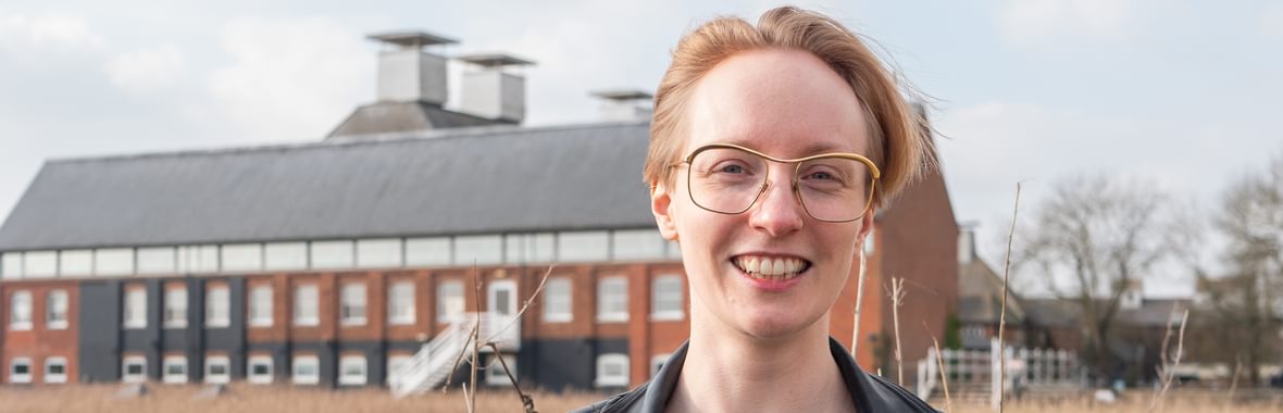woman with short, fair hair and glasses, smiling at the camera, with a maltings concert hall building in the background.