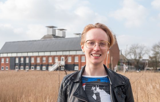 woman with short, fair hair and glasses, smiling at the camera, with a maltings concert hall building in the background.