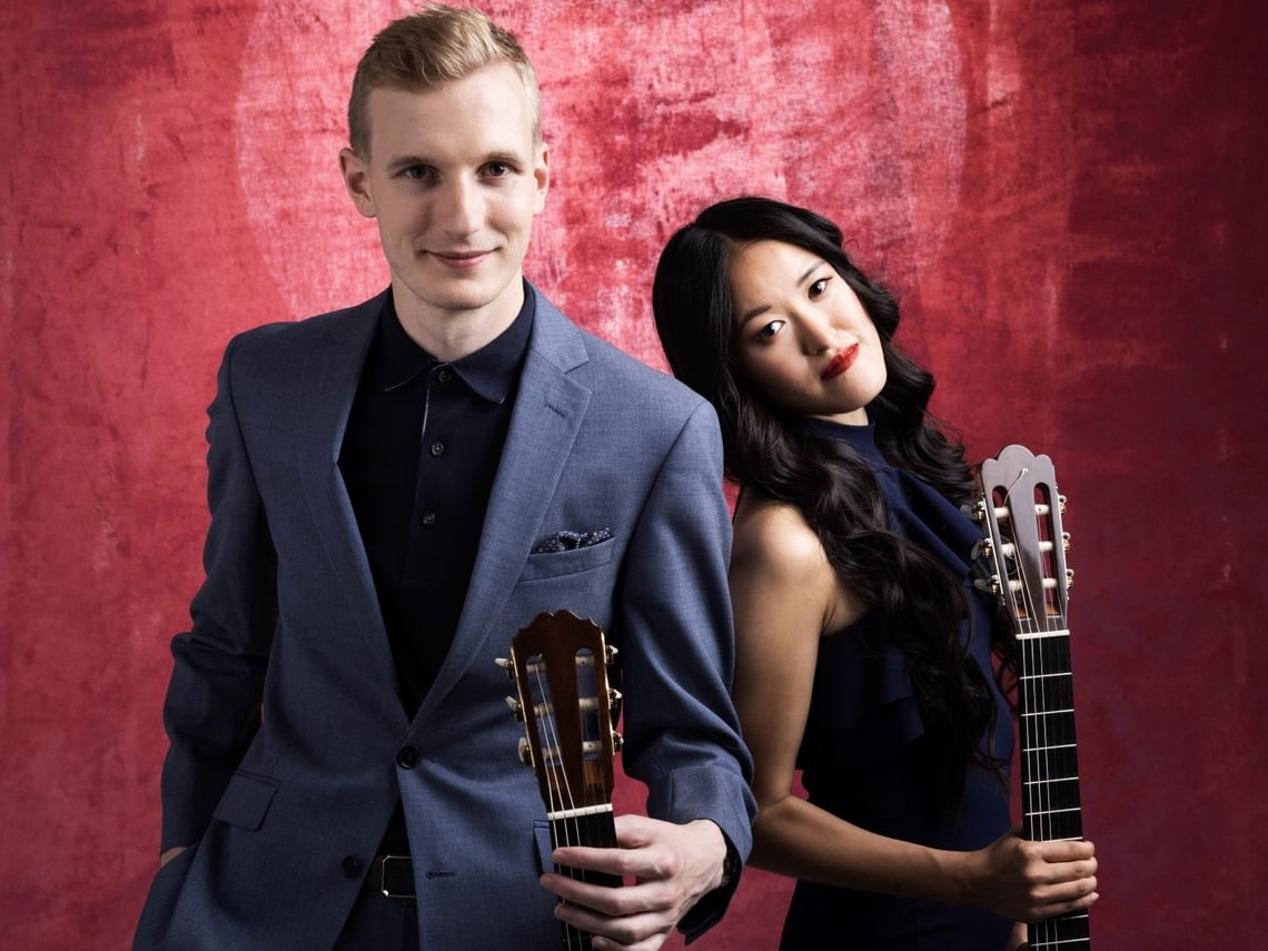 Man and woman standing close together holding guitars.