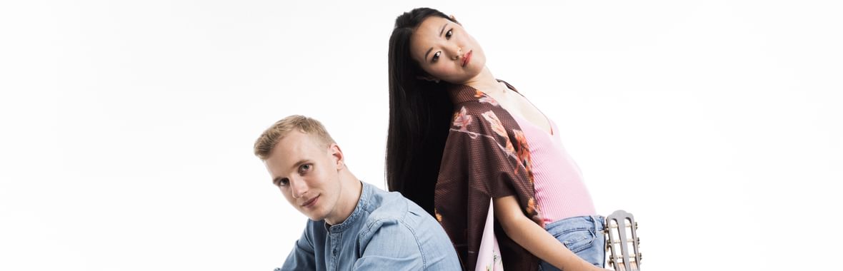 Woman with long black hair and man with short blond hair standing back to back.