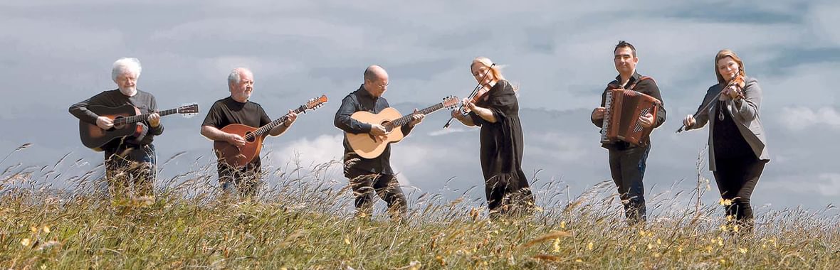 Altan musicians standing and playing instruments outside amidst long grass with a cloudy sky behind them
