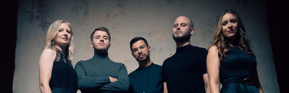 Apollo 5 standing in a group looking down at the camera against a shadowy background