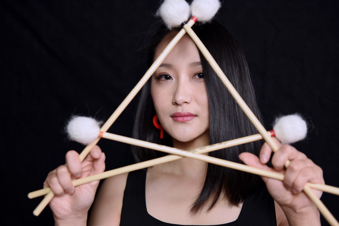 Close-up of musician Beibei Wang looking at the camera holding percussion instruments against a black background