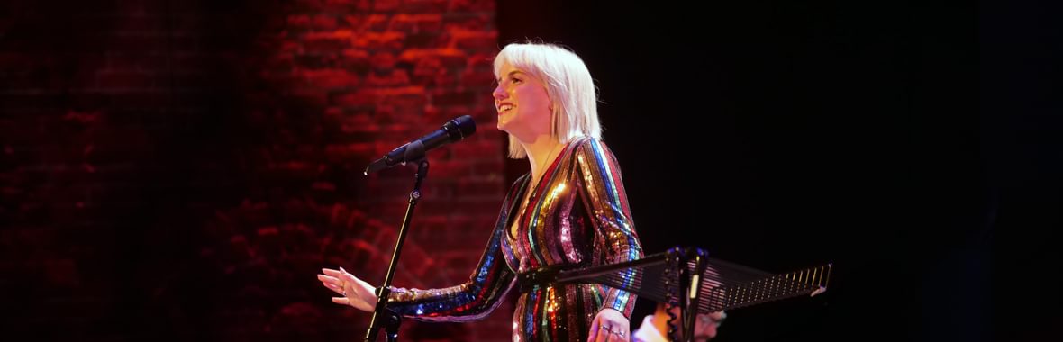 Side-on view of blonde singer performing against a dark brick background inside Snape Maltings