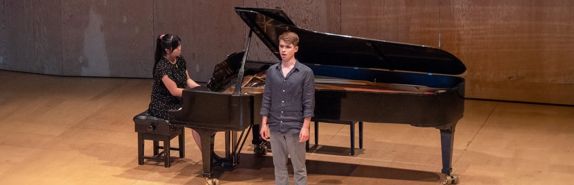 Singer standing on a wooden stage with a pianist playing a grand piano behind them