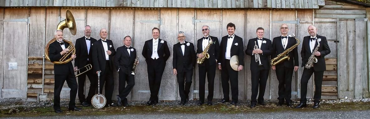 The Pasadena Roof Orchestra standing with their instruments against a wooden fence outside