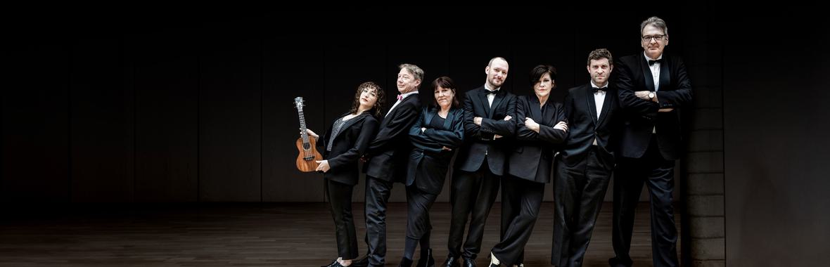 Ukulele Orchestra of Great Britain standing a in a row against a black background