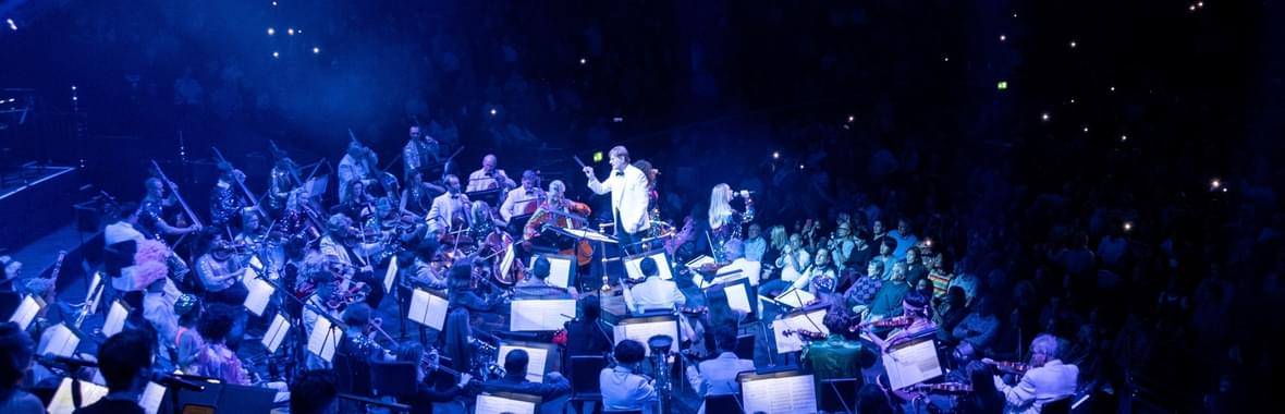 Orchestra with conductor on a blue-lit stage.