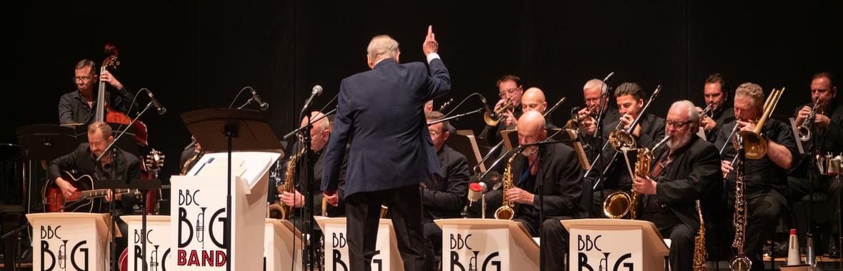 Brass band on stage with their conductor with music stands showing BBC Big Band logo.