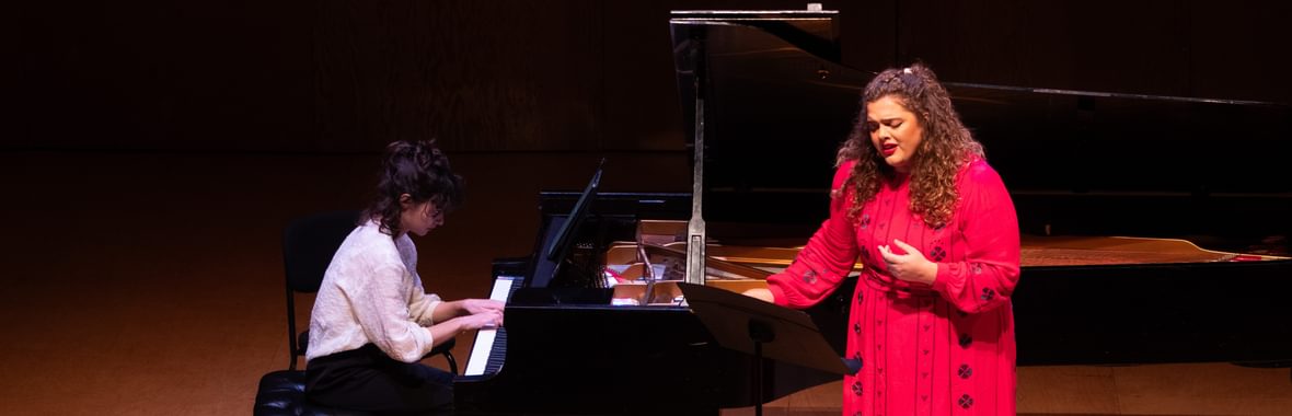 Woman in a red dress with long brown hair singing, beside a grand piano being played by a woman with dark hair wearing a white shirt.