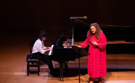 Woman in a red dress with long brown hair singing, beside a grand piano being played by a woman with dark hair wearing a white shirt.