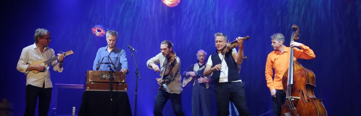 Group of men standing onstage playing instruments against a blue-lit backdrop.