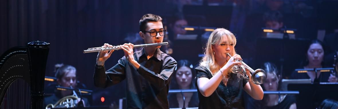 Young man with glass wearing a black shirt playing the flute and young woman with blond hair playing the trumpet, with an orchestra behind them.