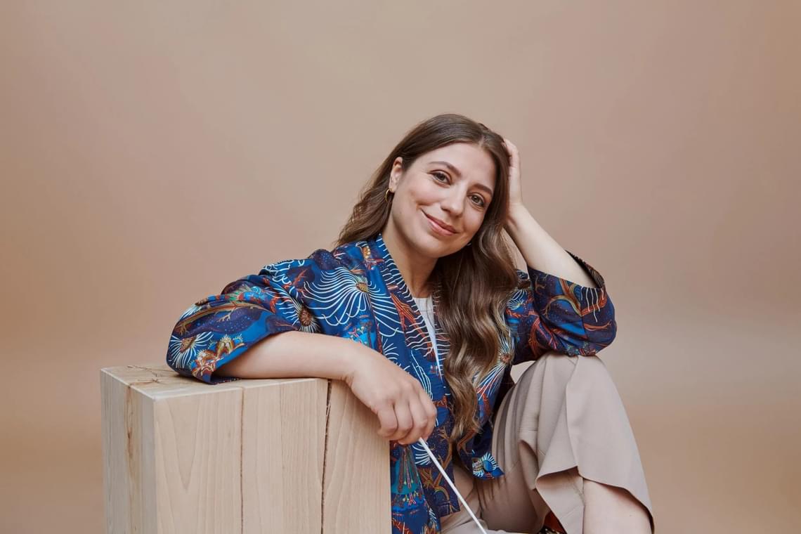 Woman with long hair wearing a patterned jacket, sitting and holding a conductor's baton and smiling at the camera.