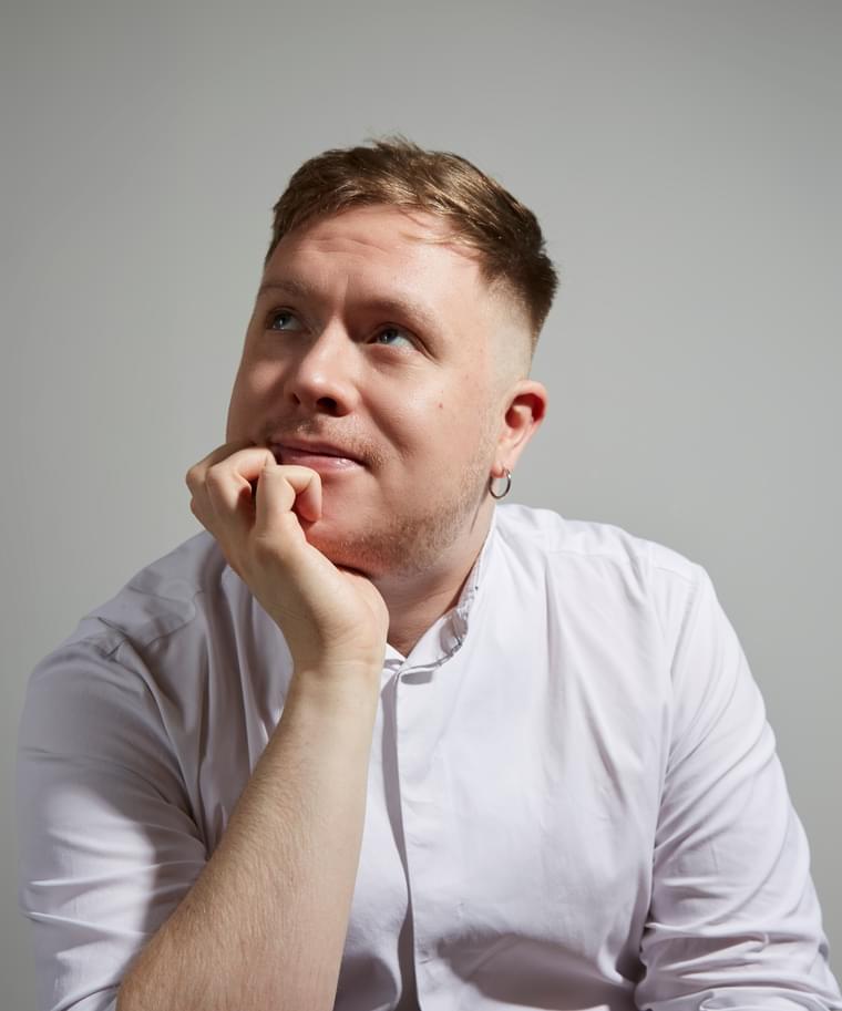 Man with cropped hair wearing a white shirt resting his chin on his hand, and looking upwards.