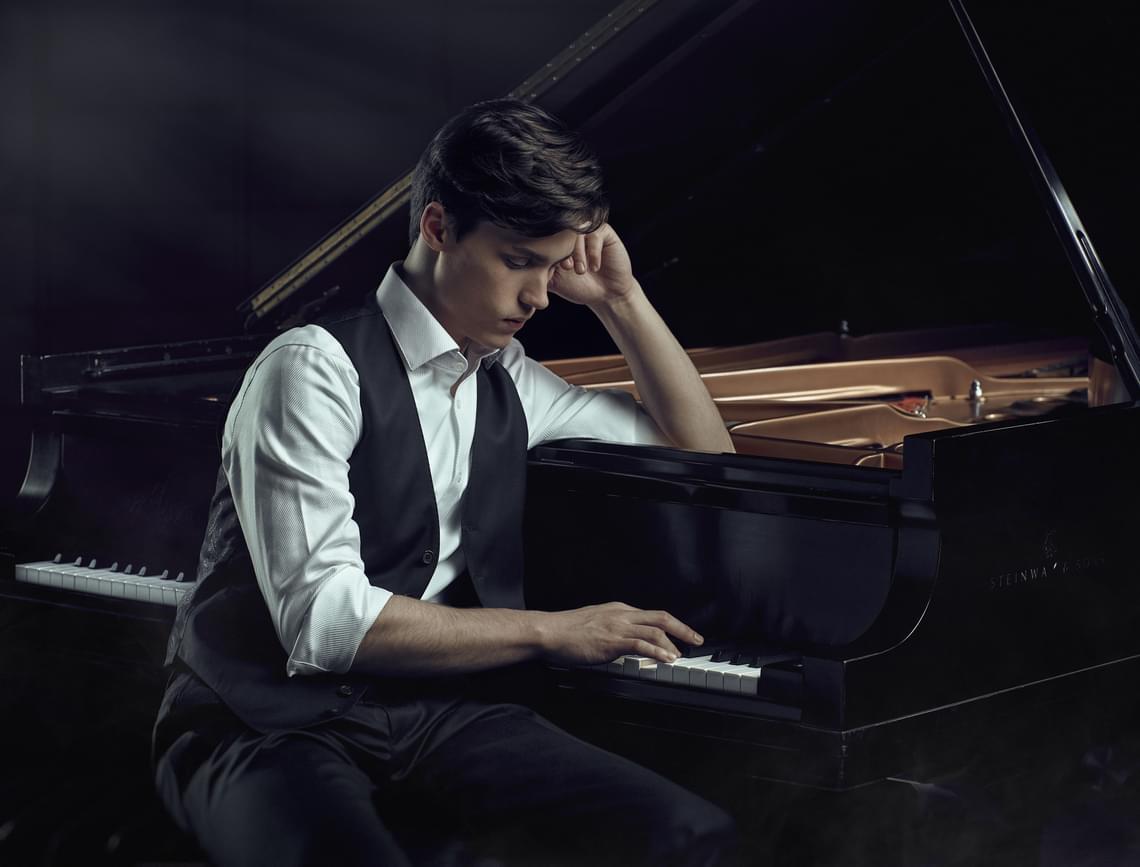 Man with short dark hair in a white shirt sitting at a piano with one hand on the keys.