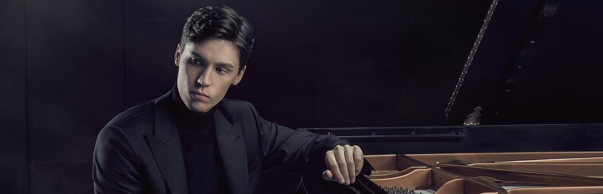 Man with short dark hair resting his arm on a grand piano and looking away from the camera.