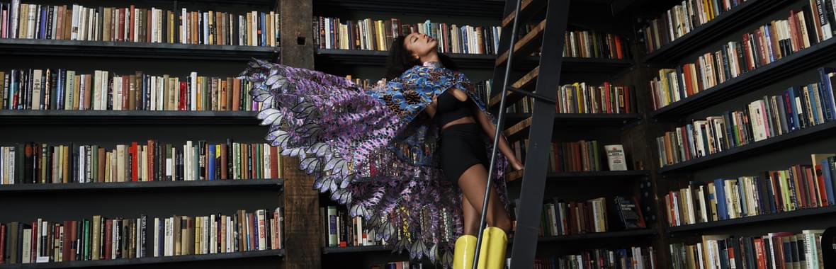 Women surrounded by bookshelves standing on a ladder with a patterned cape and bright yellow boots.