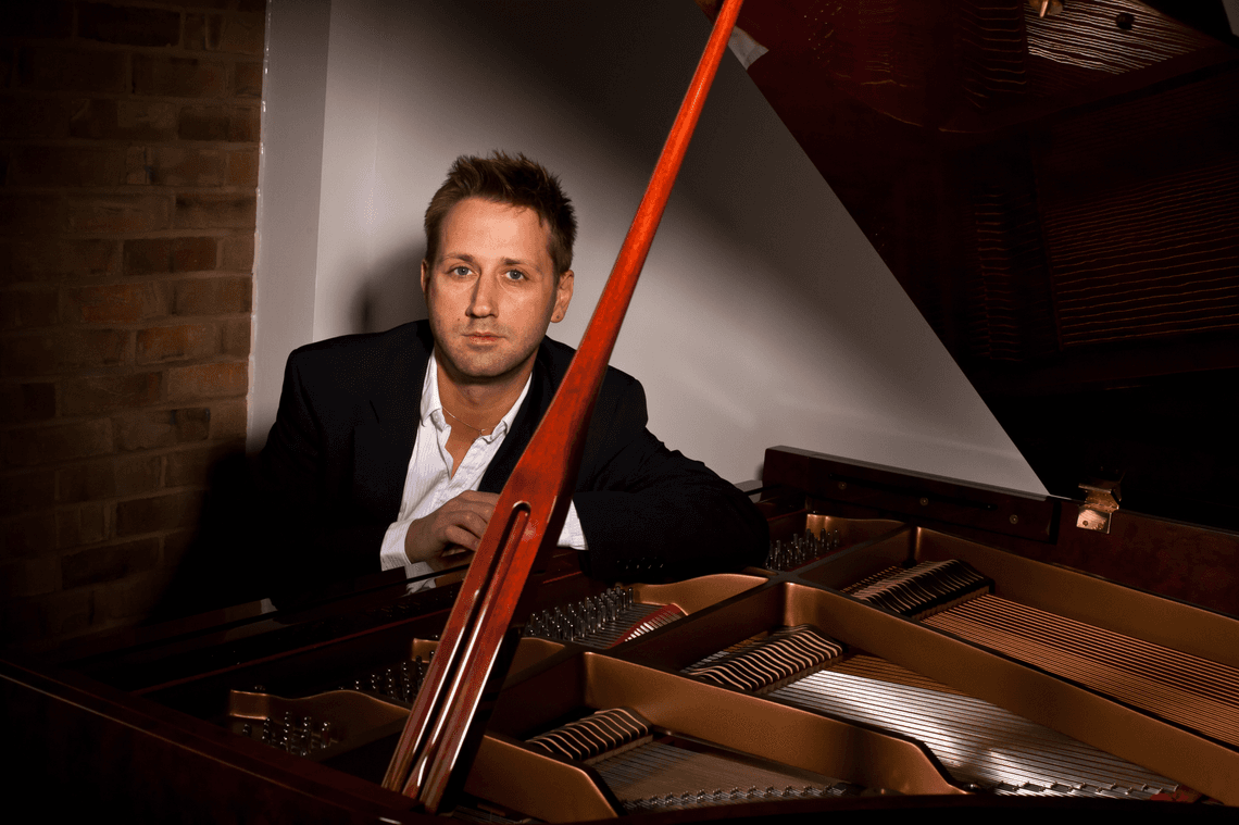 Man with short hair wearing a shirt and jacket sitting at the piano and looking at the camera.