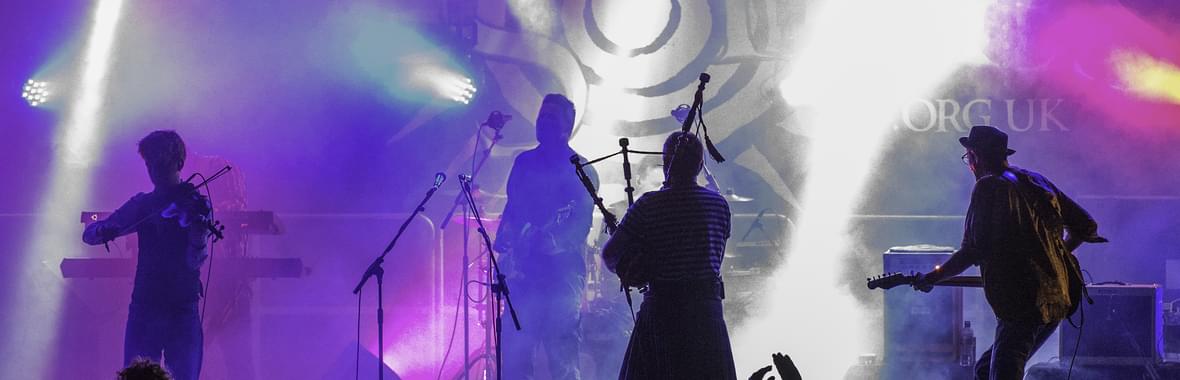 Group on stage playing violin and bagpipes silhouetted against purple and blue lighting.