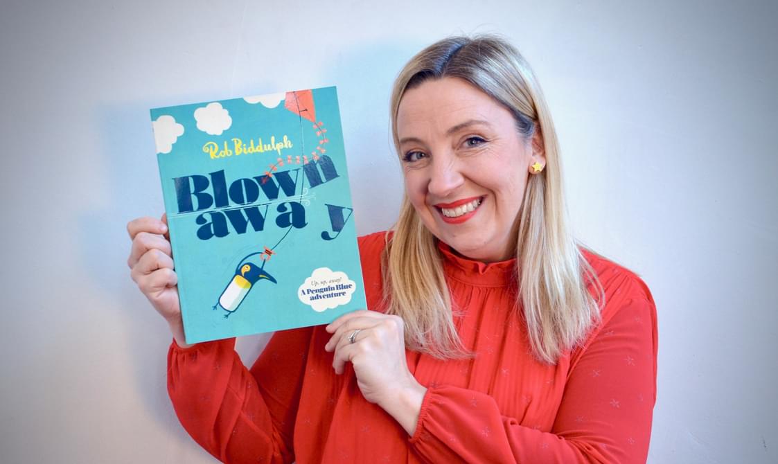 Woman with blond hair and red jumper holding up a book and smiling.