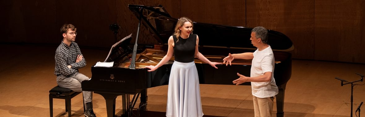 Man sitting at a grand piano and woman standing beside it, both listening to another man who is gesturing and speaking to them.